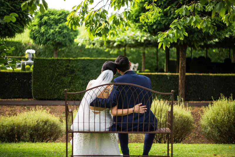 Les mariés assis de dos sur un banc en fer forgé, dans un jardin.