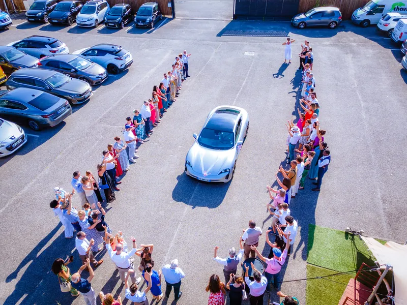 Vue de drone : la voiture des mariés avance entre deux haies d’invités.