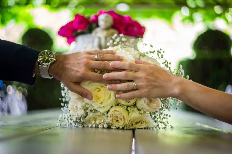 Les mains des mariés posées sur le bouquet, alliances visibles.