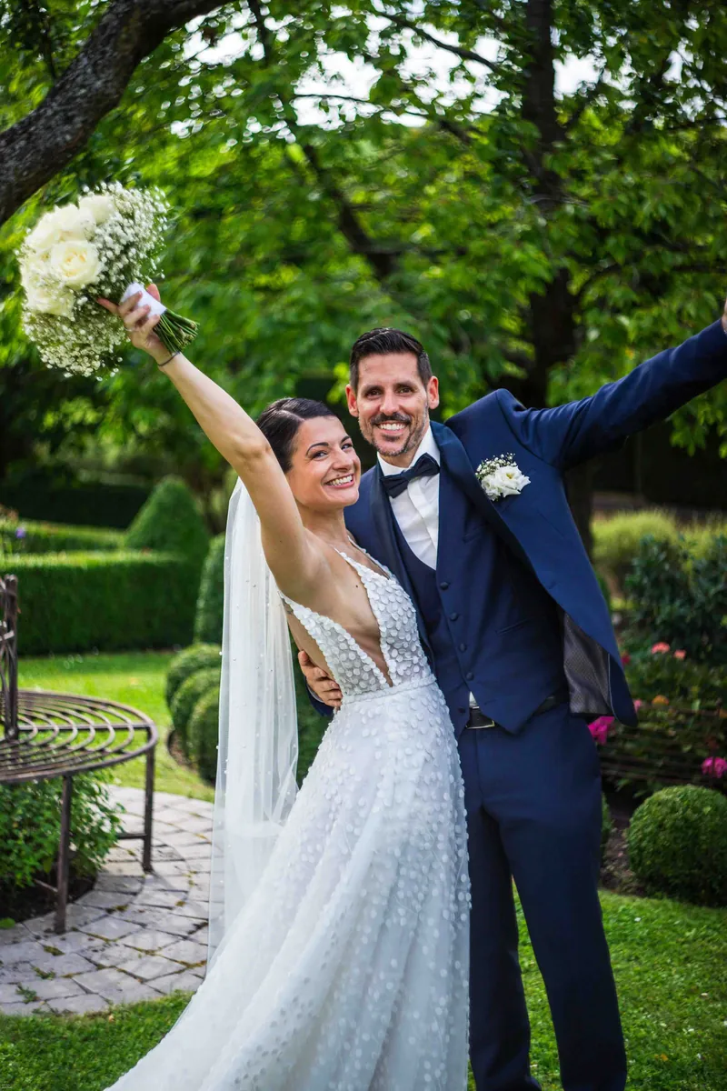 La mariée lève son bouquet de roses blanches, le marié ouvre grand les bras, dans un jardin à la française.