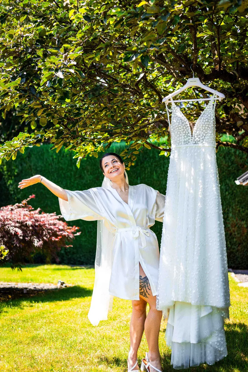 Préparatifs : la mariée en peignoir à côté de sa robe suspendue à un arbre.