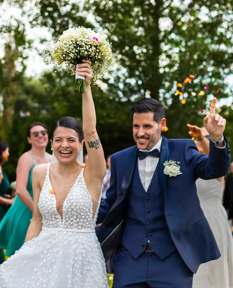 Sortie de cérémonie sous les pétales : la mariée lève son bouquet en riant.